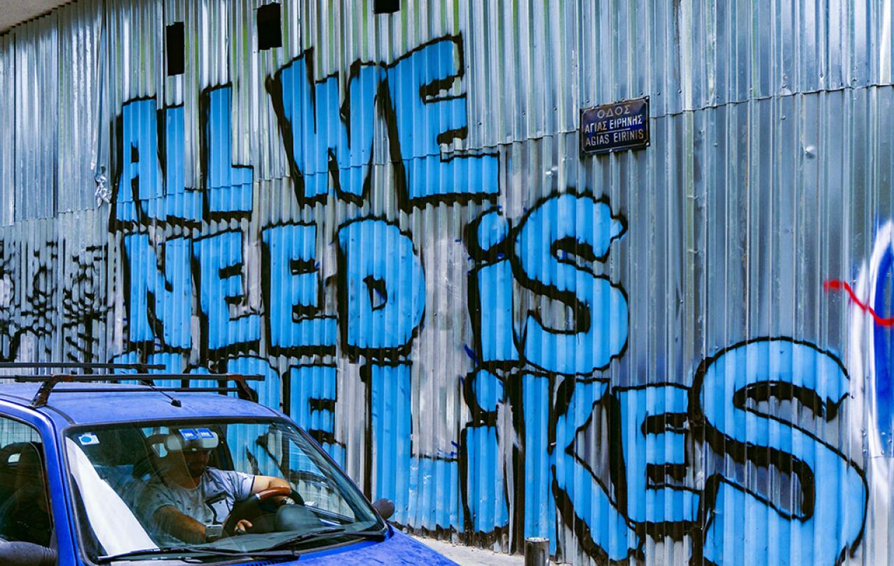A corrugated sheet iron wall graffited in large blue letters that say 'All we need is more likes.'