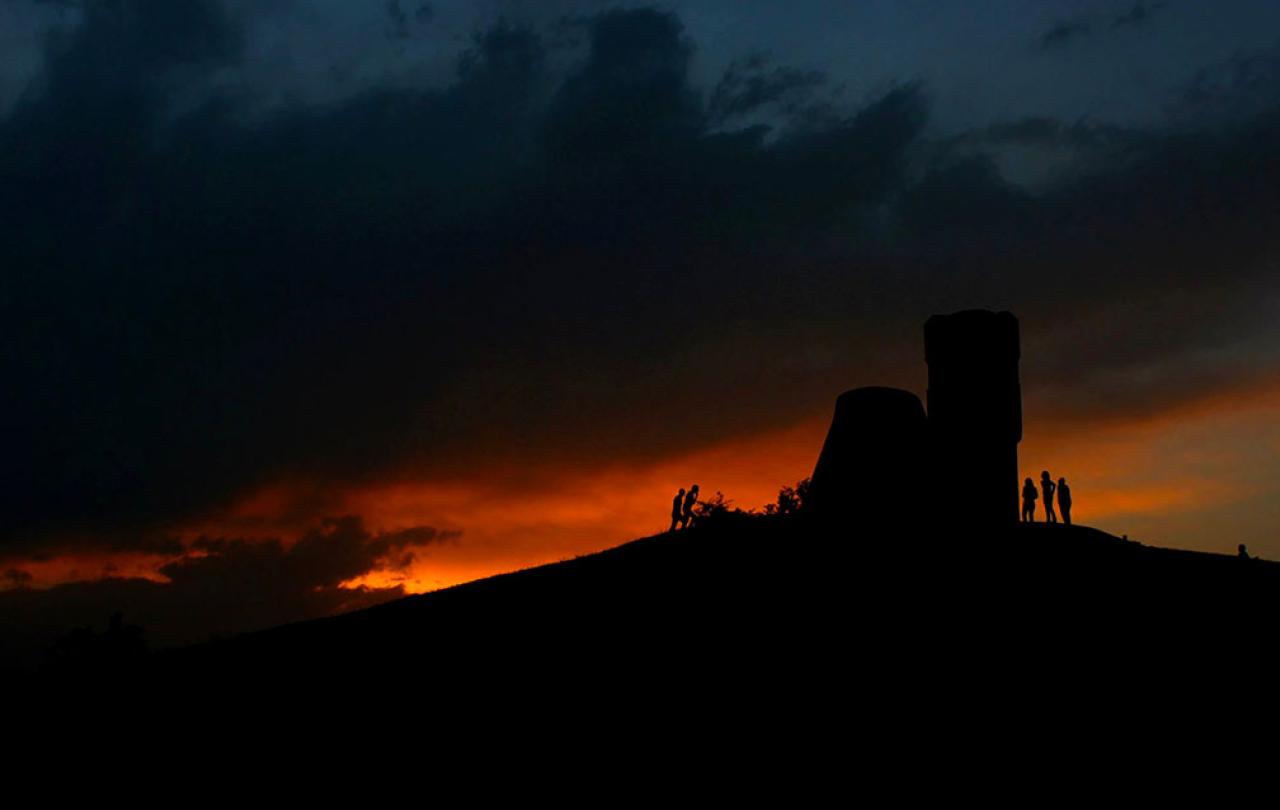 A sunset dramatically silhouette's a ruined tower and people at its base.