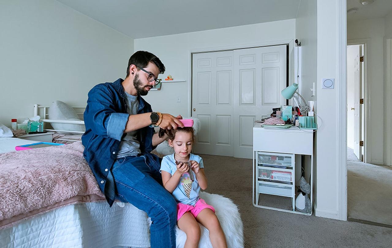 A father sits on a bed and fixes the hair of his daughter standing in front of him