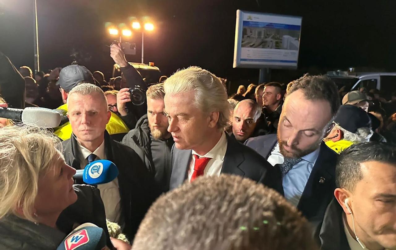 A politician in a suit stands amid a scrum of reporters holding microphones