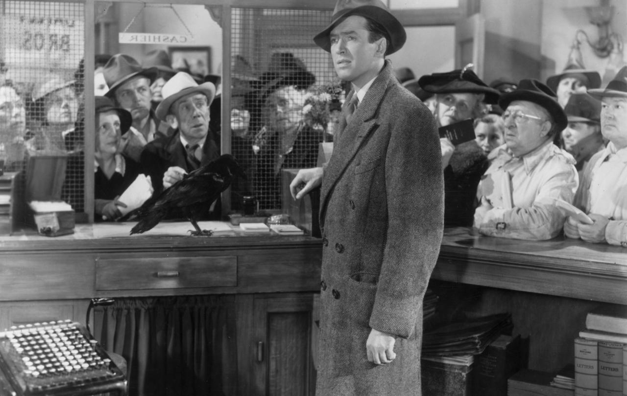 A man stands one side of a bank counter while others, on the other side, look hopefully at him.