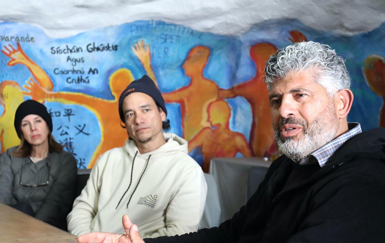 Two people down a table turn and listen to someone closer talk, against a wall mural.