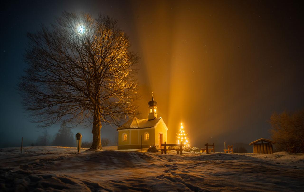A nocturnal snow-covered scene of a tree, chapel and Christmas tree casting shadows.