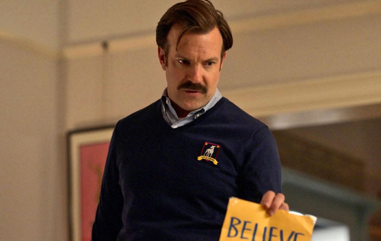 A man in a blue jumper holds a yellow sign reading 'believe'.