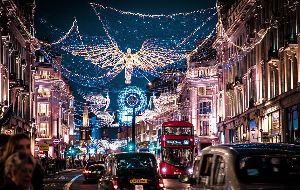 A shopping street is crowded by taxi cabs and buses while above it a Christmas illumination of an angel hangs over all.