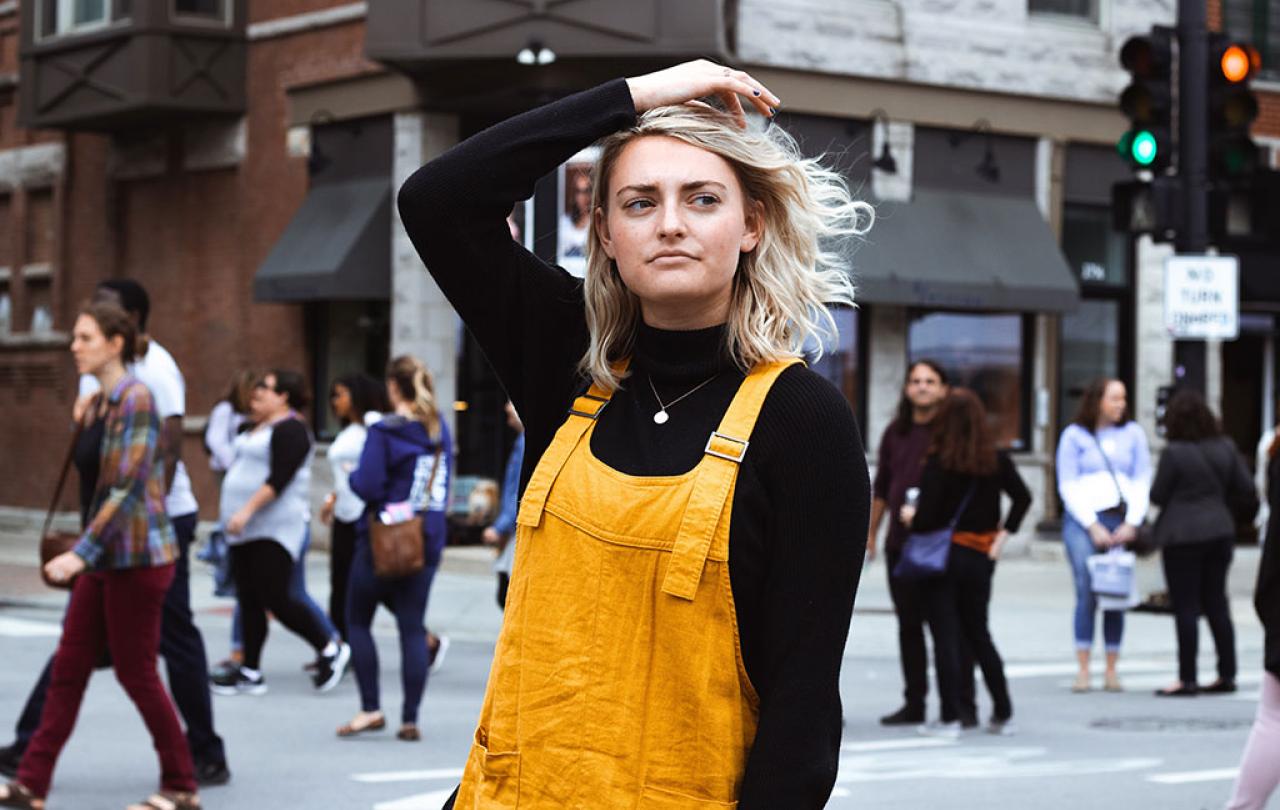 A woman stops in her stride down a street and pensively runs her hand through her hair as she looks to the side.