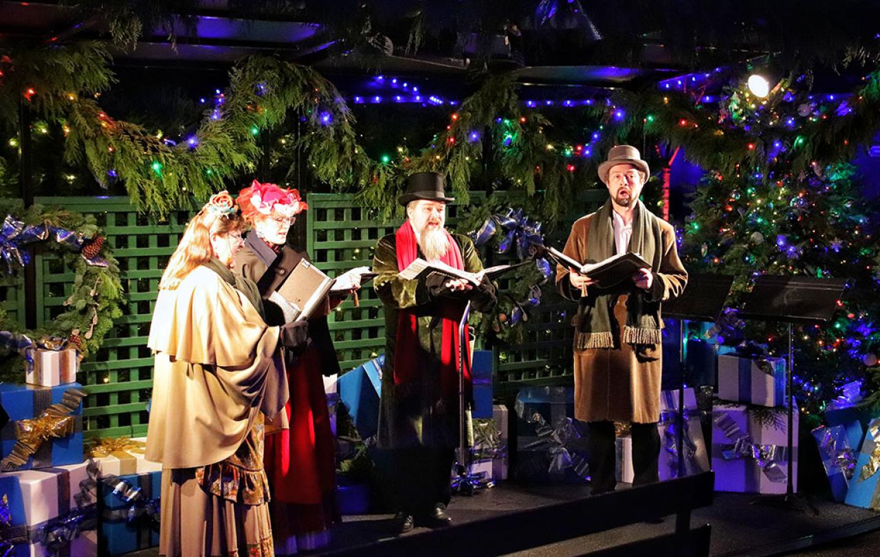 Dressed in Victorian clothes, a group of carol singers stand and sing amid Christmas foliage.