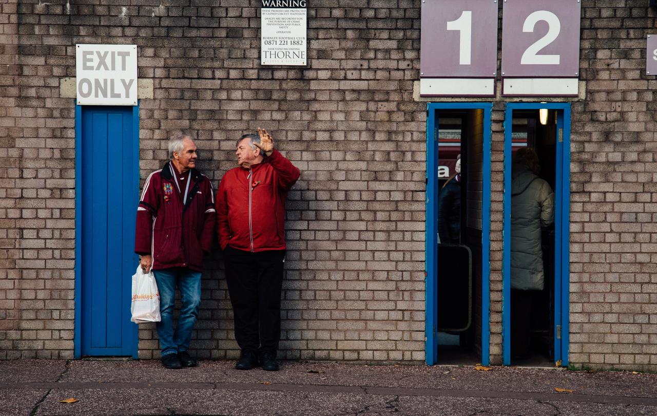 Two older men lean agains the wall of a football club's entrance, next to thin open doorways.