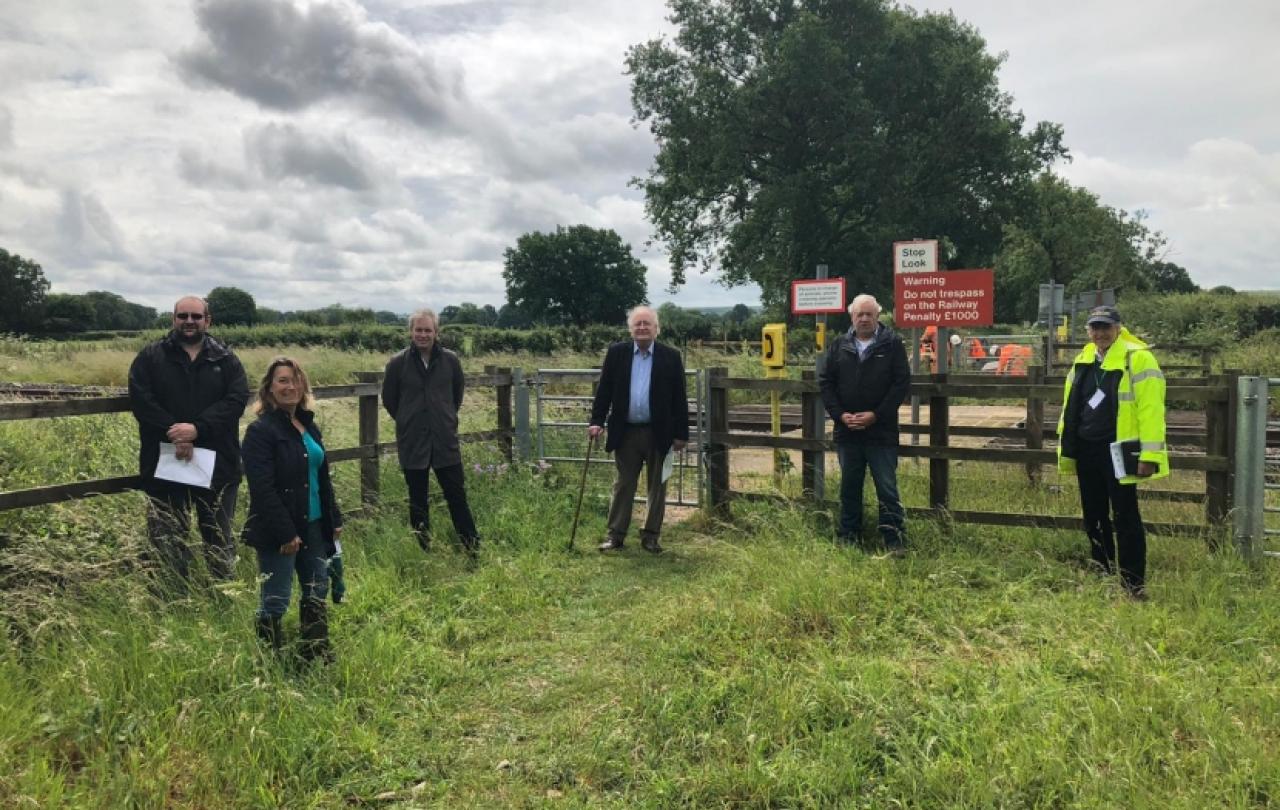 A group of people stand in a field by a fence and a railway footcrossing.