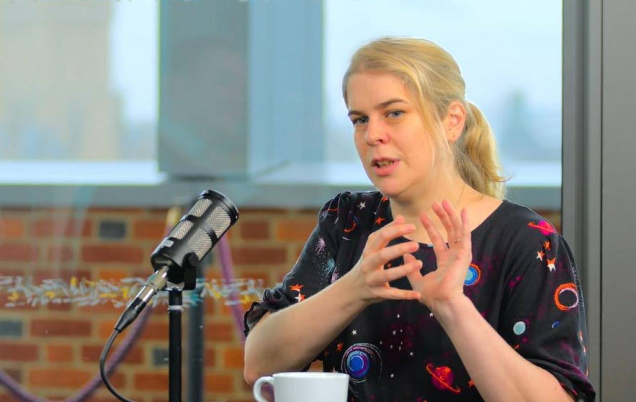 a woman sits at a table, with a mic in front of her, talking and holding her hands together in front of her.