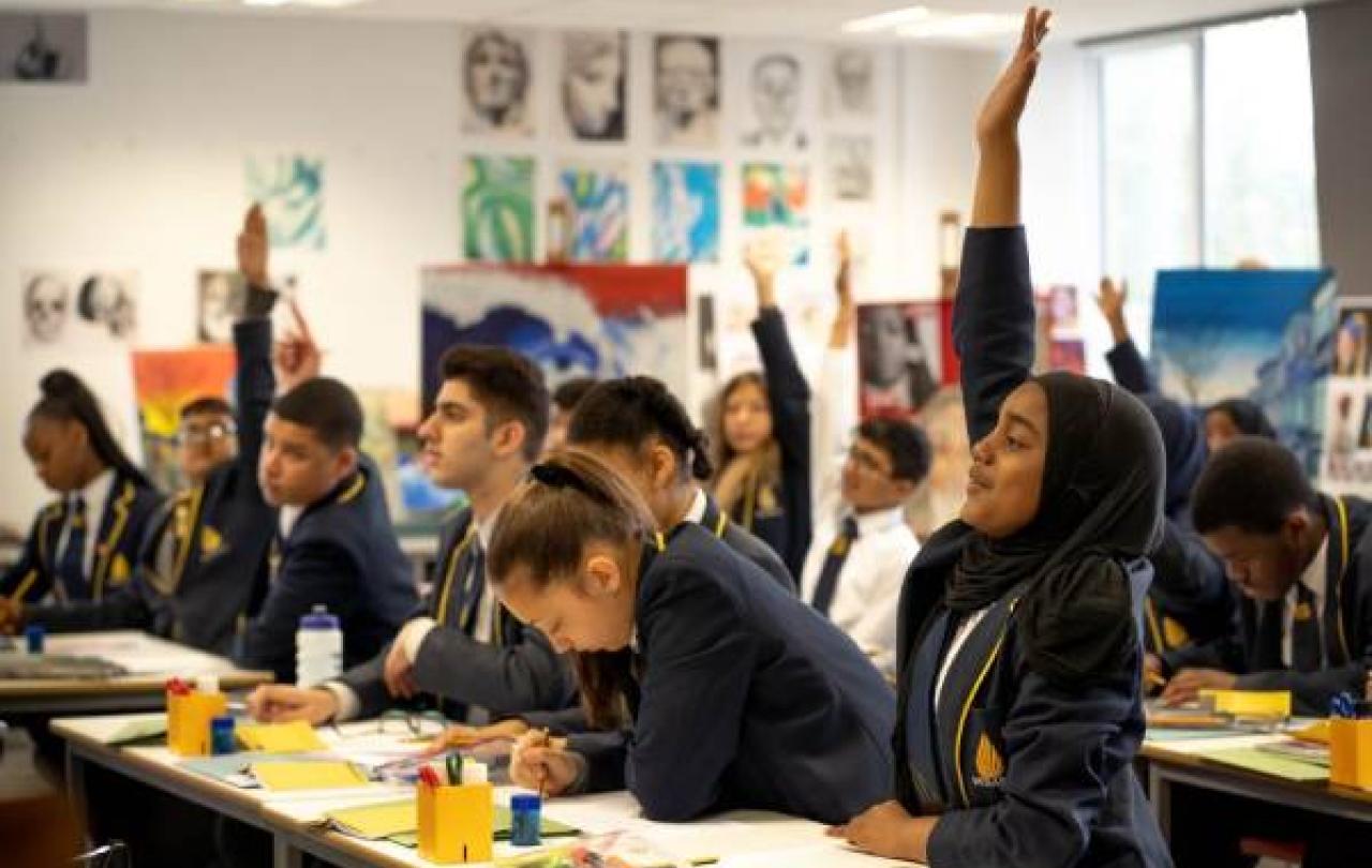 school pupils sit at desk, some with a hand raised.