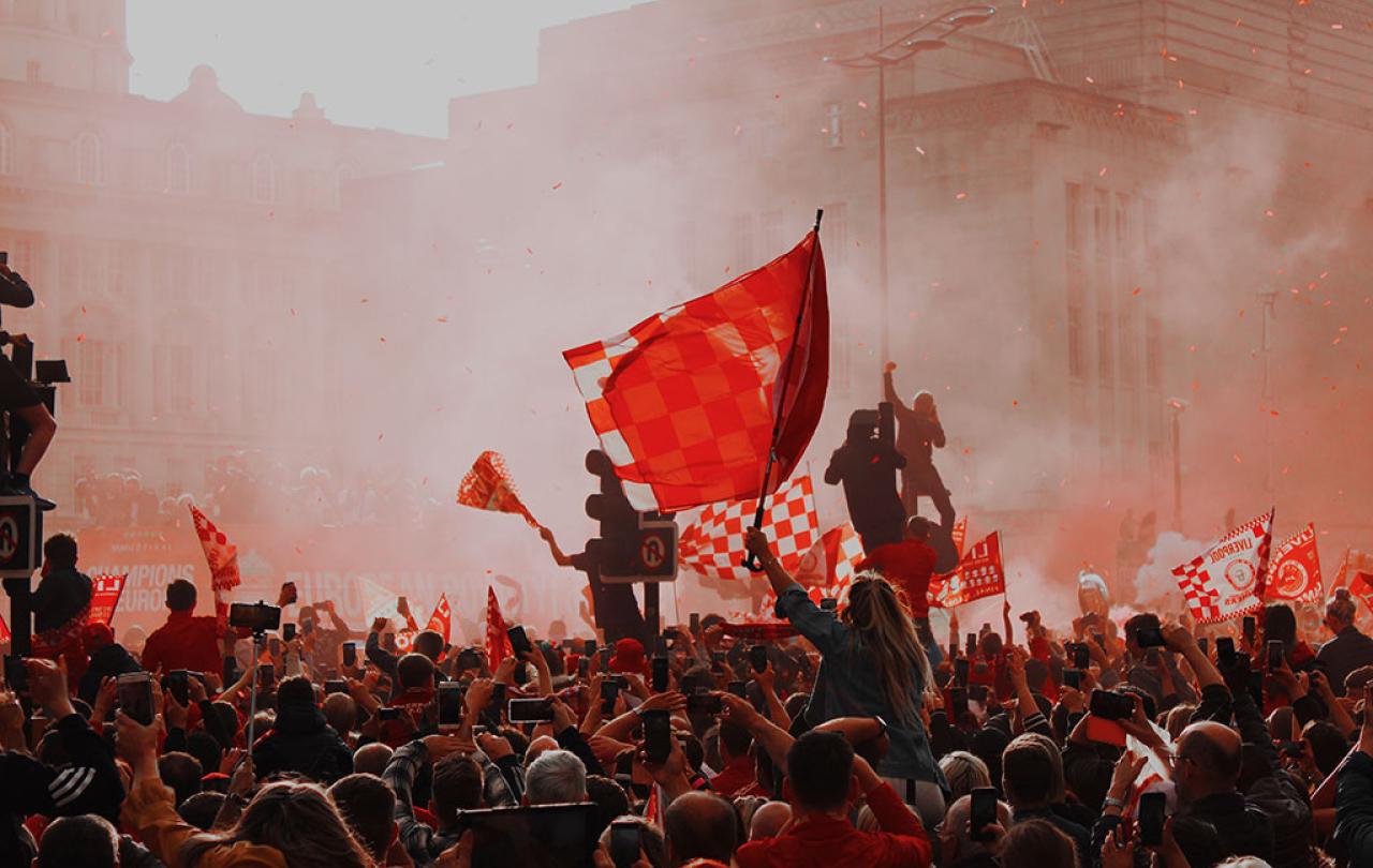 Silouhetted by red flare smoke, celebrating footballs wave red flags.