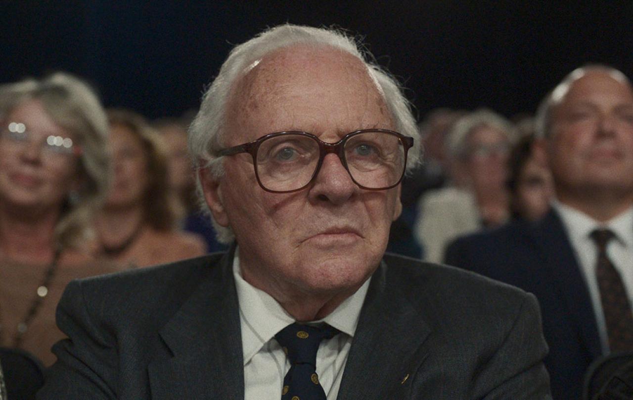 An old man wearing a suit and tie sits in a TV audience as people stand around him.