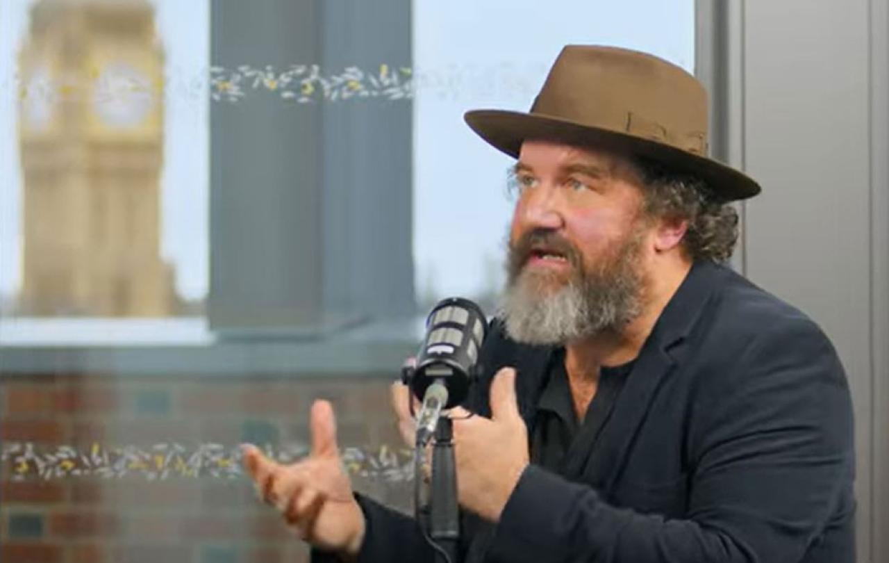 A man wearing a hat sits at a table talking and raises both hands in front of himself to gesture