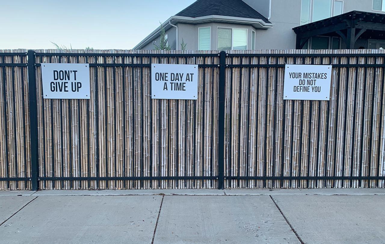 Three signs attached to a fence read: Don't Give Up, One Day At A Time, and Your Mistakes Don't Define You.
