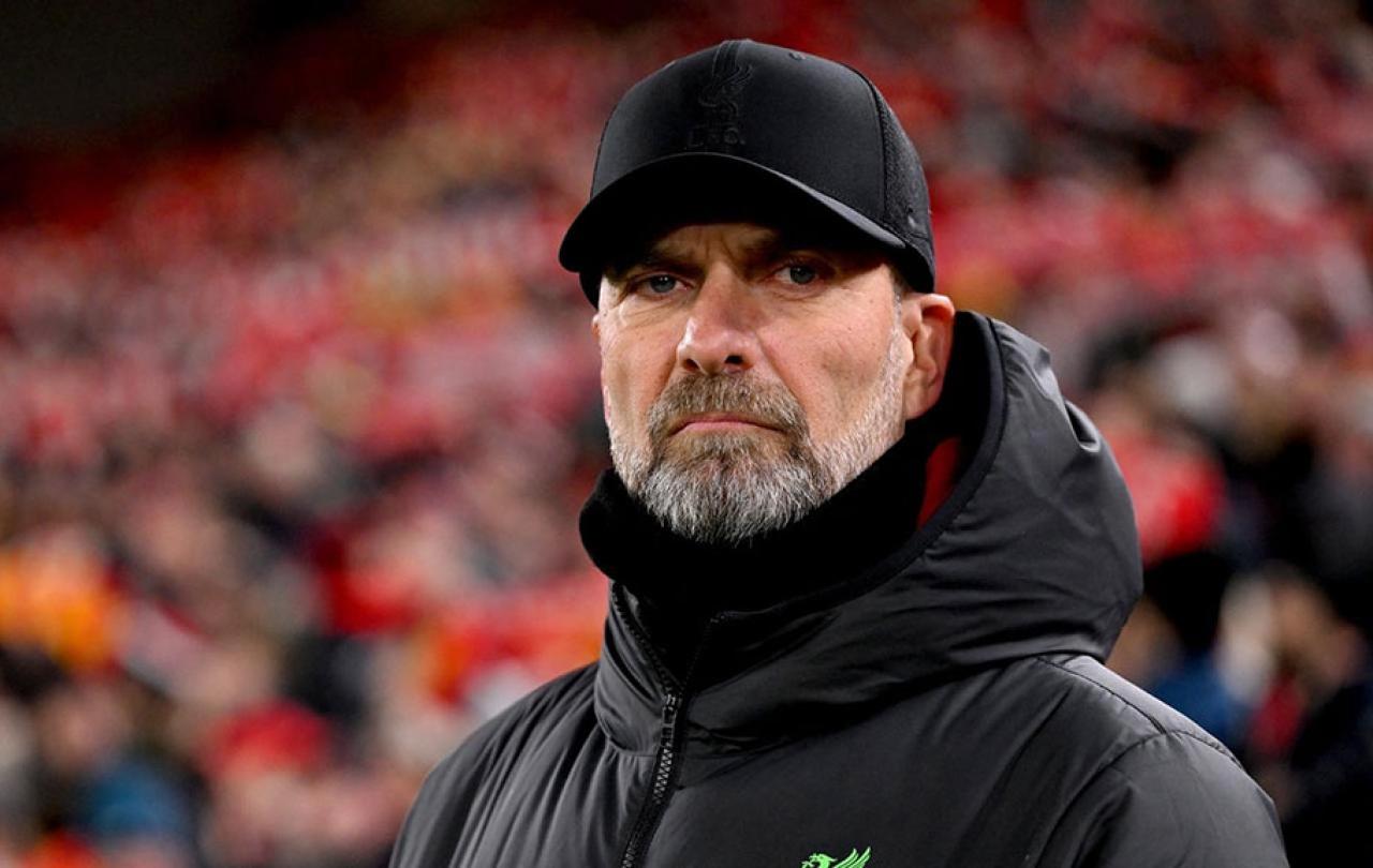 A football manager stands on the touchline and stares hard, dressed in a black coat and hat.