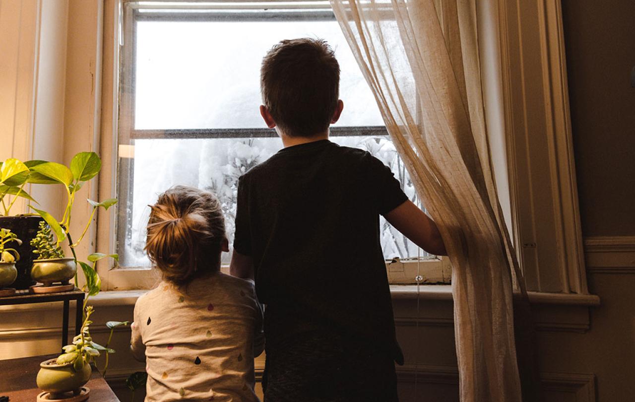 Two small chidlren push a curtain aside to stare out the window at snow.