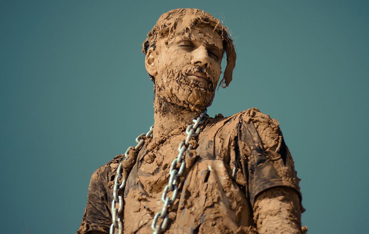 A man covered in dried and caked mud stands and looks to the side, a steel chain is draped from his shoulders.