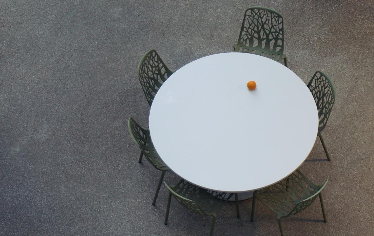 a round table with empty chairs is seen from above. An orange is the only item on it.