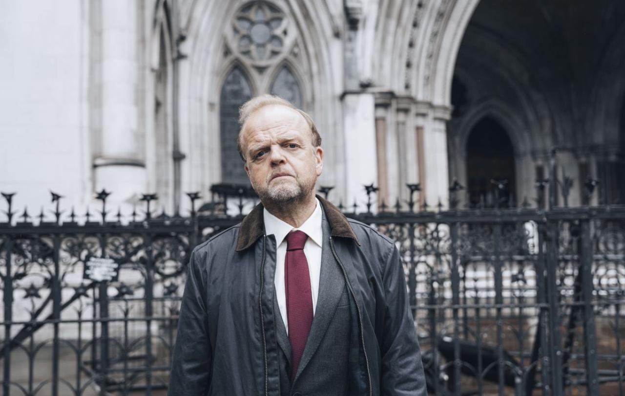 A man, dressed in a suit and anarak, stands in front of a law court.