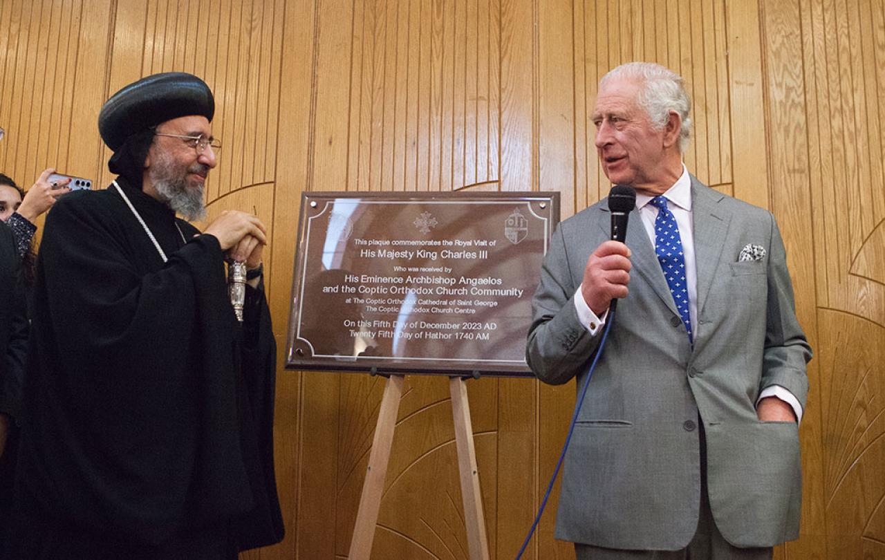 An archbishop wearing a black hat and robes stands next to a new building's plaque, while King Charles, wearing a suit stands the other side holding a mic.