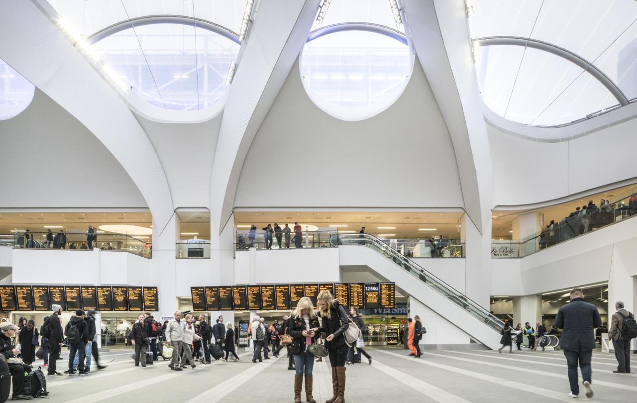 A airy and light station concourse in which people stand and look at a long set of travel information screen.