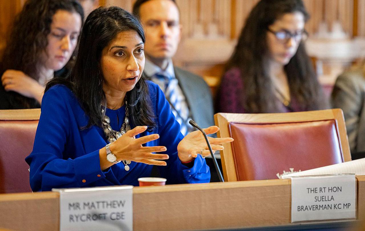 A woman dressed in a blue suit sits at a table talking and gesticulating with her hands.