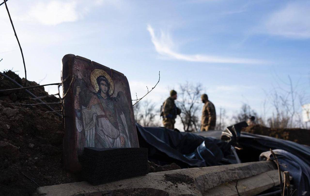 A religious icon is propped on the side of a trench, a soldier can be seen further down the trench.