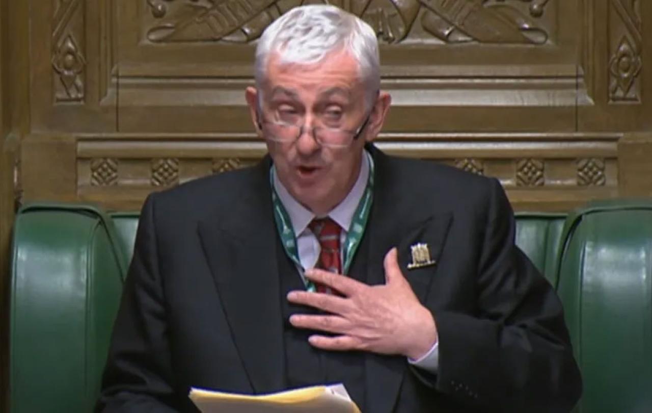 A robed and seated man, in a speaker's chair talks and gesticulates.