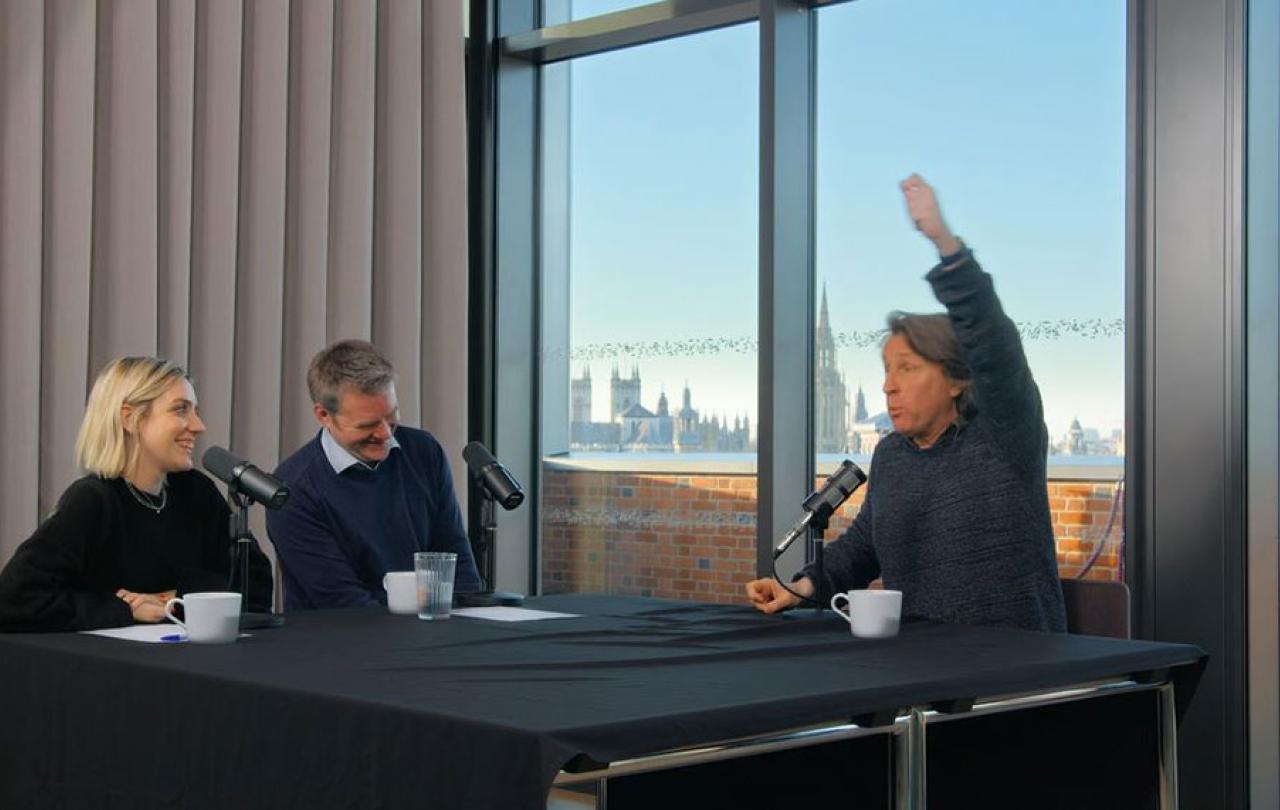 A sitting man raises one hand above his head to emphasise a point, while two other people looking on are laughing.