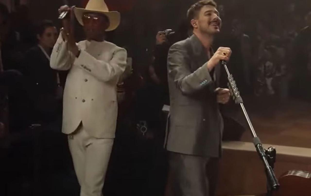 Two singers peform together. One in a white suit and stetson claps their hands. The other tilts the mic stand.