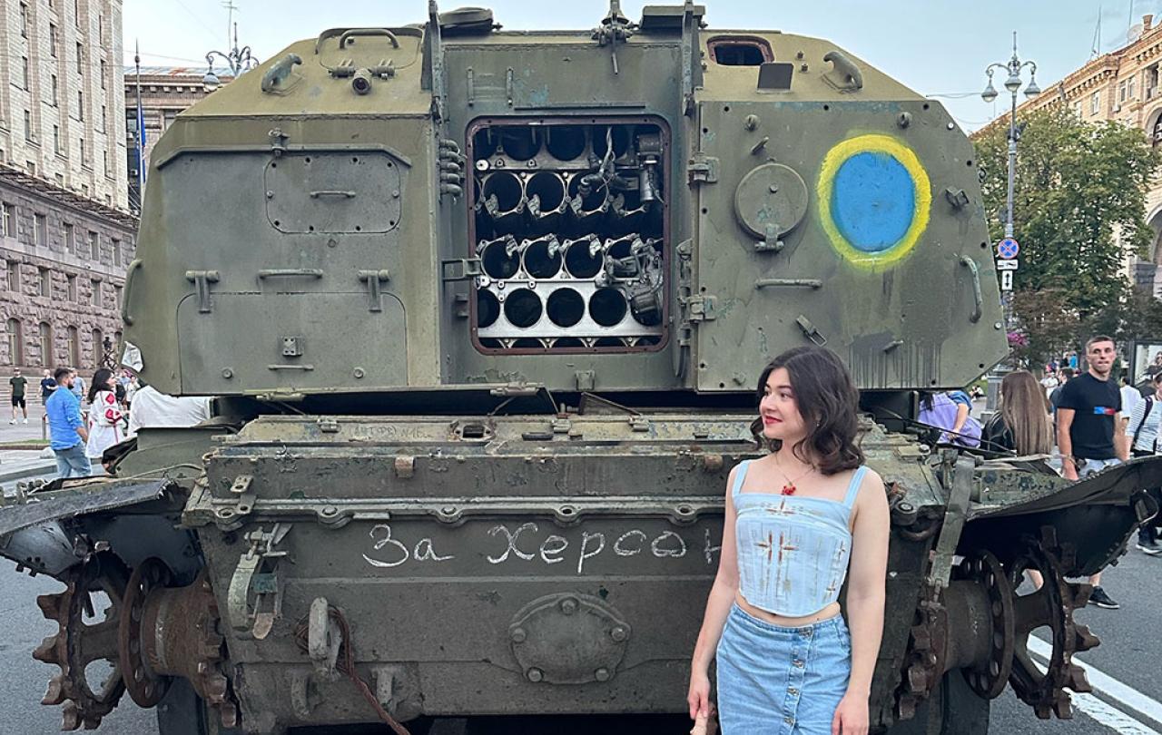 A woman stands at the back of an armoured military vehicle, the door of which is open.