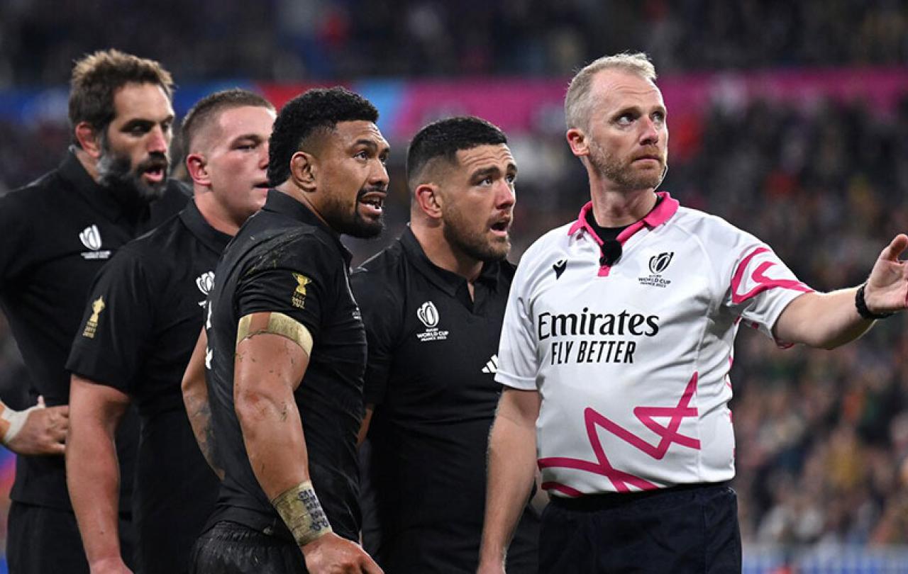 Four rugby players stand and watch beside a referee gesturing with his arm.