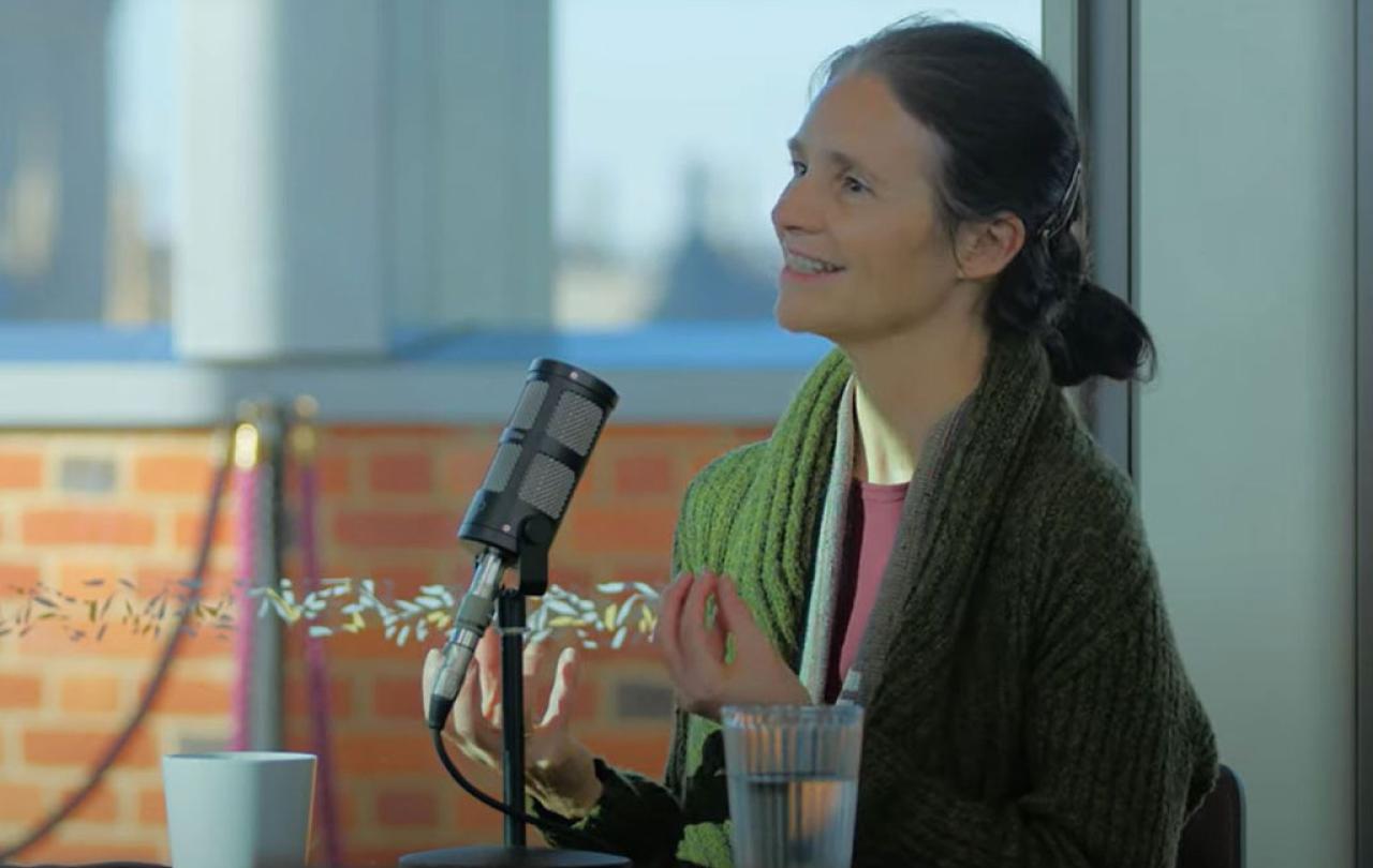 a woman sits at a table, with a mic in front of her, talking and holding her hands out in front of her.