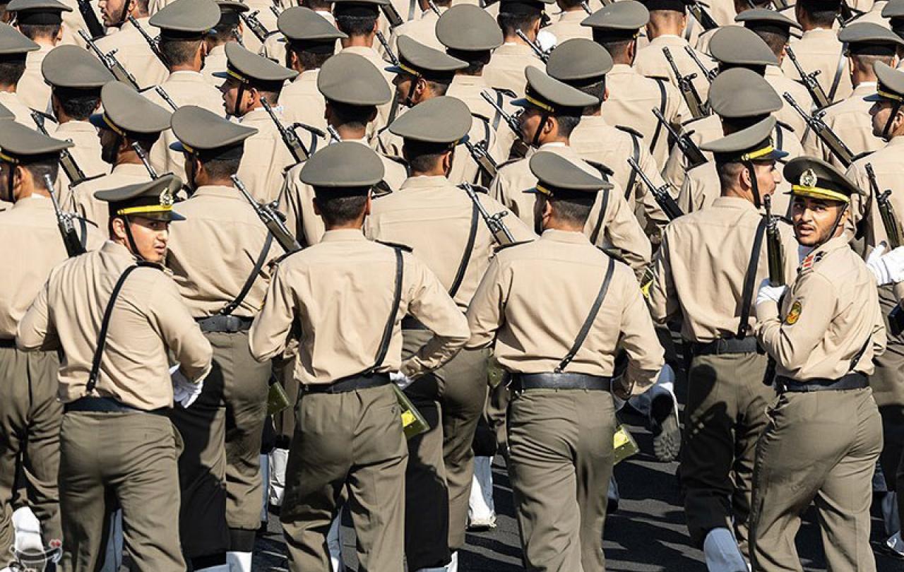 Rows of soldiers march away from the camera, two in the back row turn their heads back.