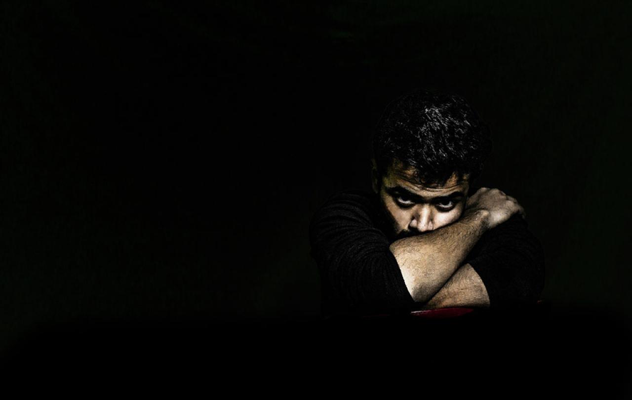 In a darkened room, a man's angry face is lit as he rests on arms folded tightly around it.