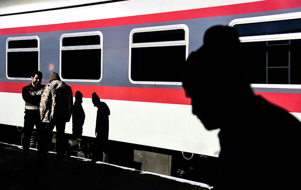 On a sun-lit railway platform, two men talk casting shadows on a waiting red and white train. Another shadow beside them is that of a man silouheted in the foreground