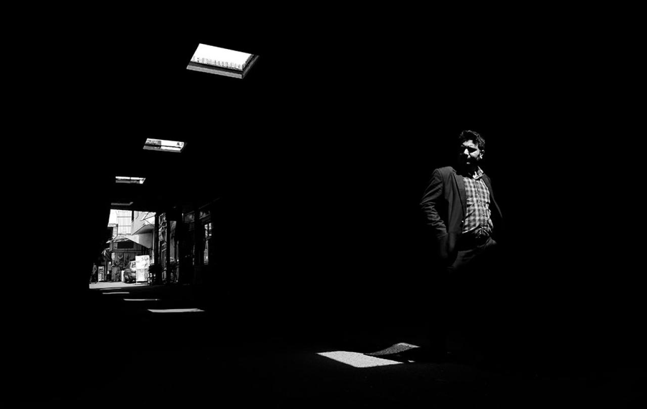 A man walks through a dark alley, looking to one side, illuminated only by roof lights.