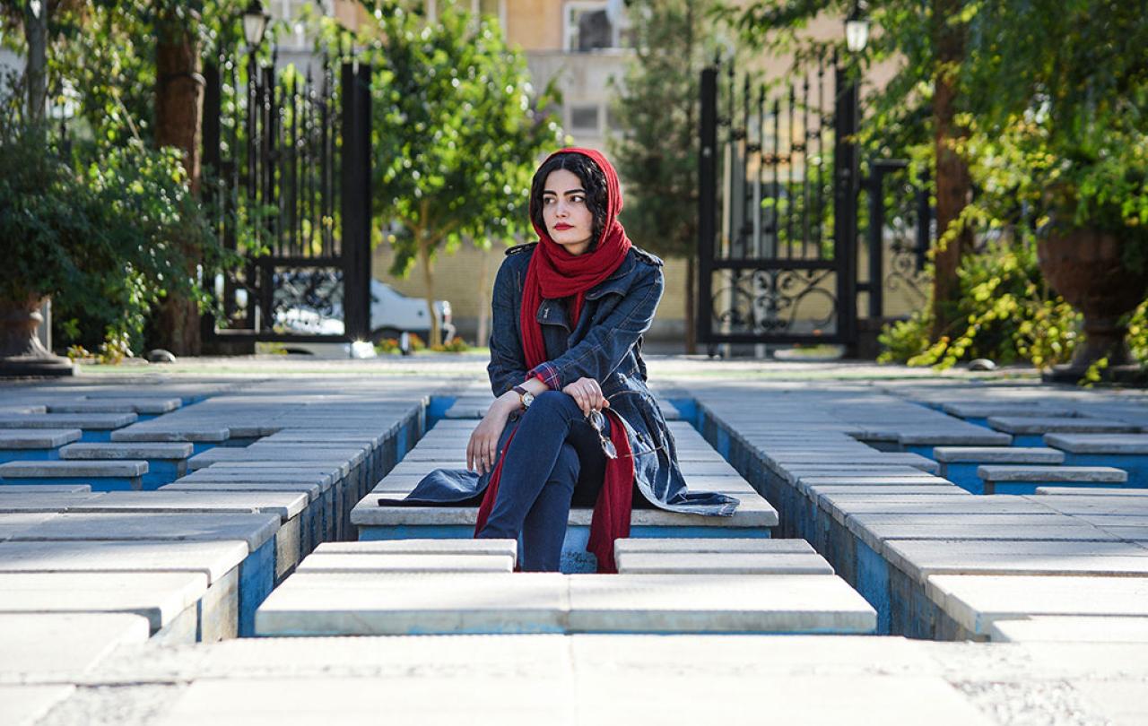 A woman wearing a headscarf sits, looking pensive, amid a grid of concrete seats.