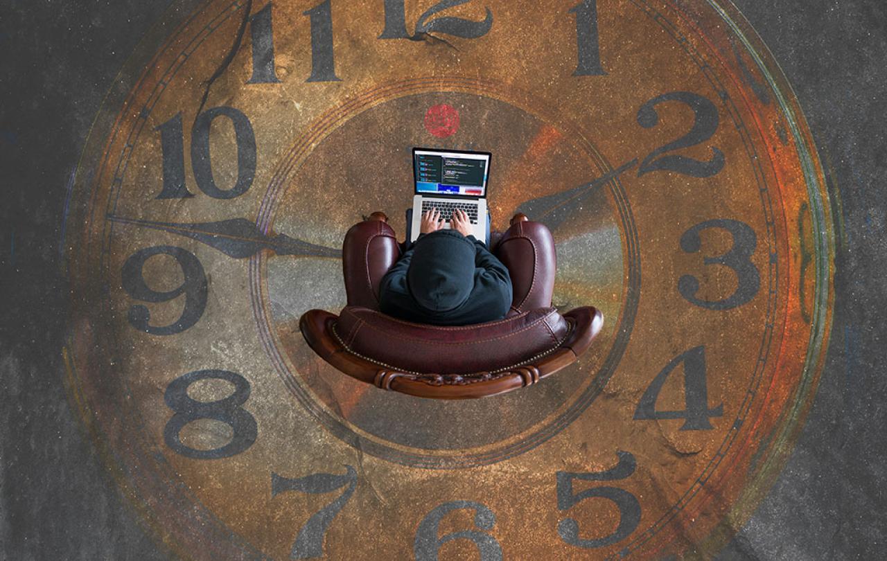 Looking straight down on someone sitting in an armchair working on a laptop. They are surrounded by clock numerals and hands on the floor.