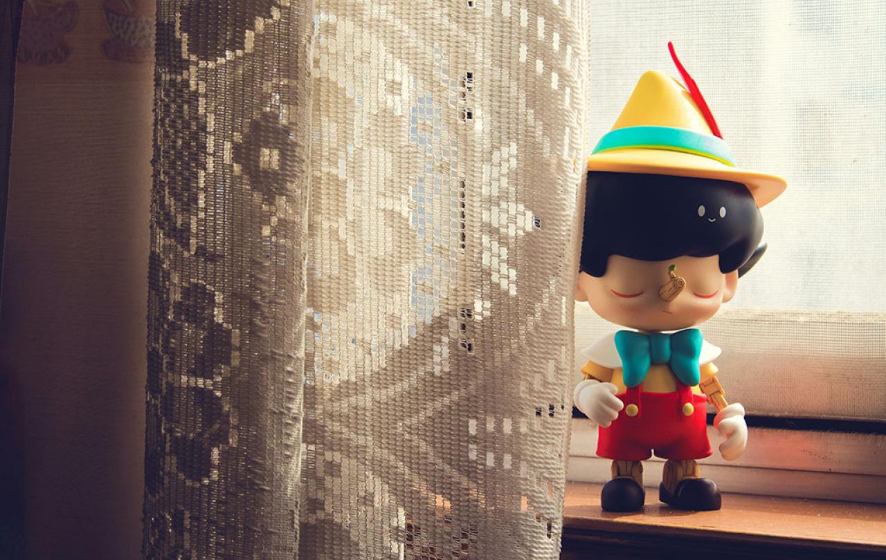 A Pinnochio figure stands on a window sill beside some net curtains.