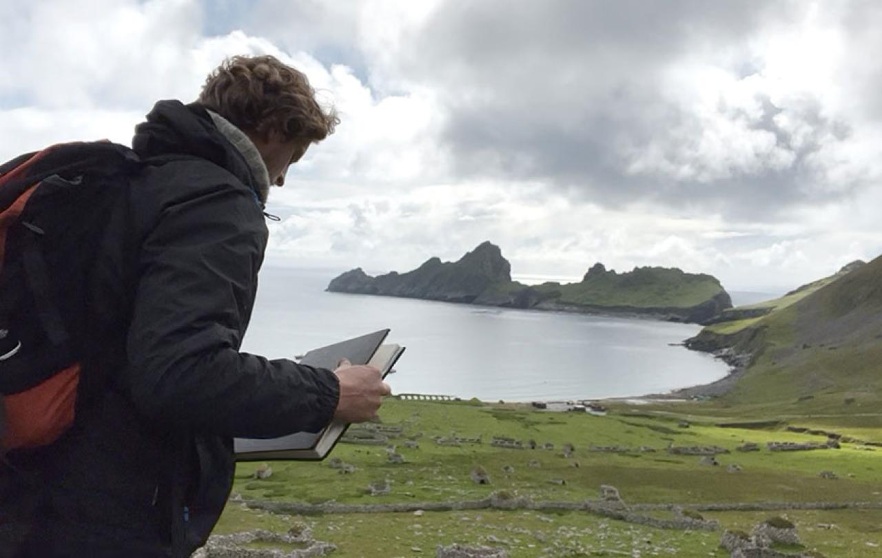 An artist holds a sketchbook while standing overlooking a deserted village by a bay, sided by jagged cliffs.