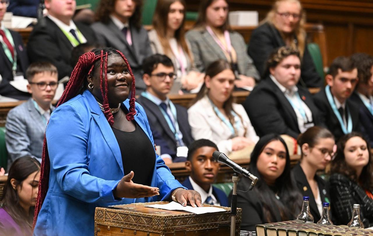 A young woman in a blue suit stands at a wooden box in a parliamentary debating chamber looking upward while speaking.