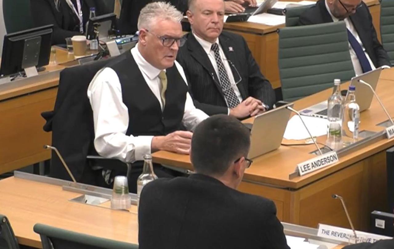 A man wearing a waist coats sits at a desk and ask a questions of a panel of people with their backs facing us.