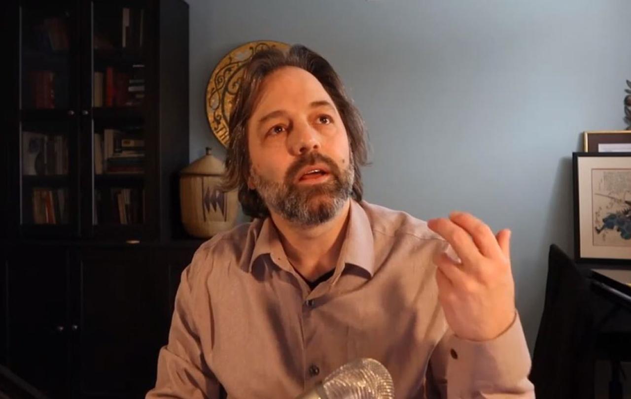 A man sits at a desk and looks up and gestures with his hand.