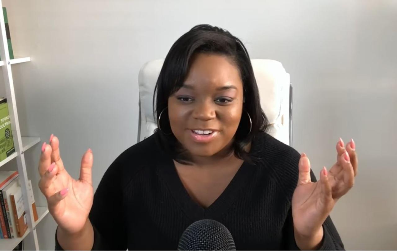 A woman sits and speaks into a desktop microphone and holds both hands up and out expressively.