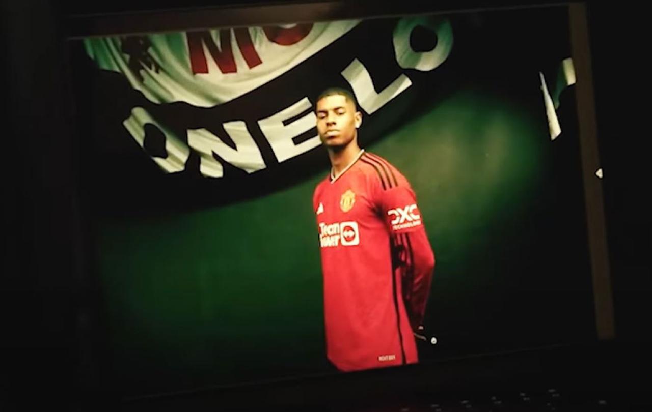 A professional footballer wearign a red top stands proudly beneath a white and black banner.