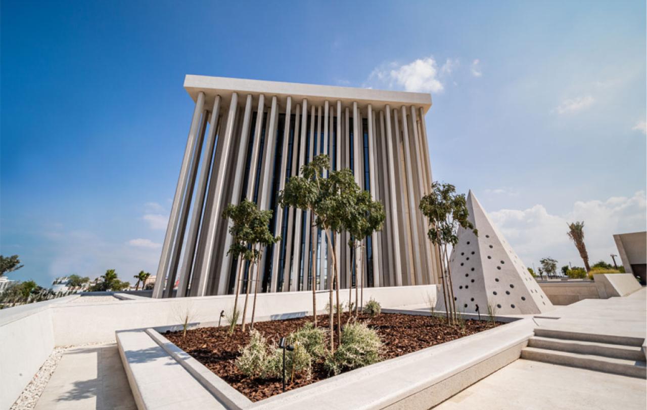 A large, modern cubiod building with a series of thin external pillars is seen from low down against a sunny blue sky 