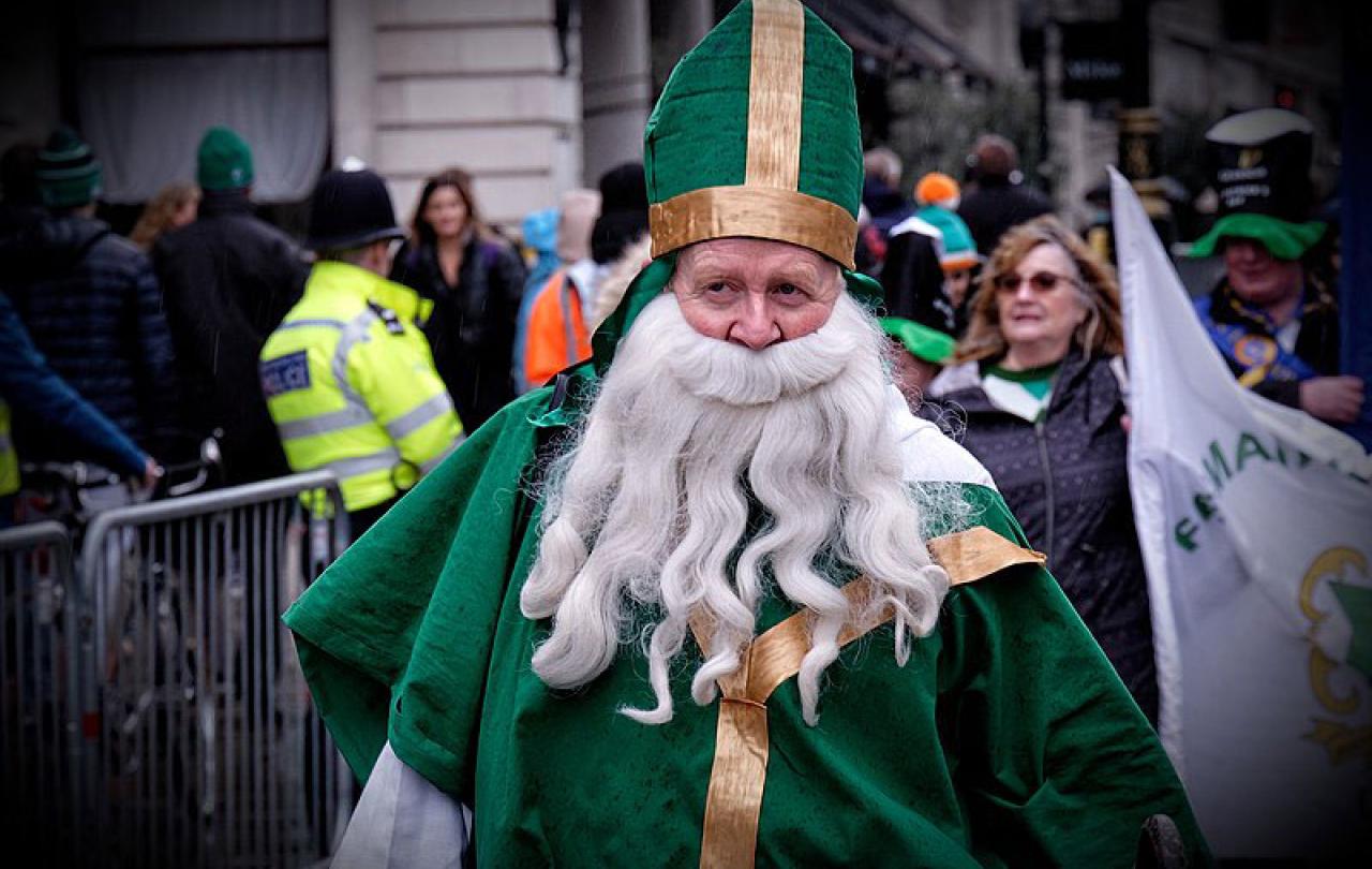 A parade particpant dressed as a bishop in green vestments with a false beard walks down a street.