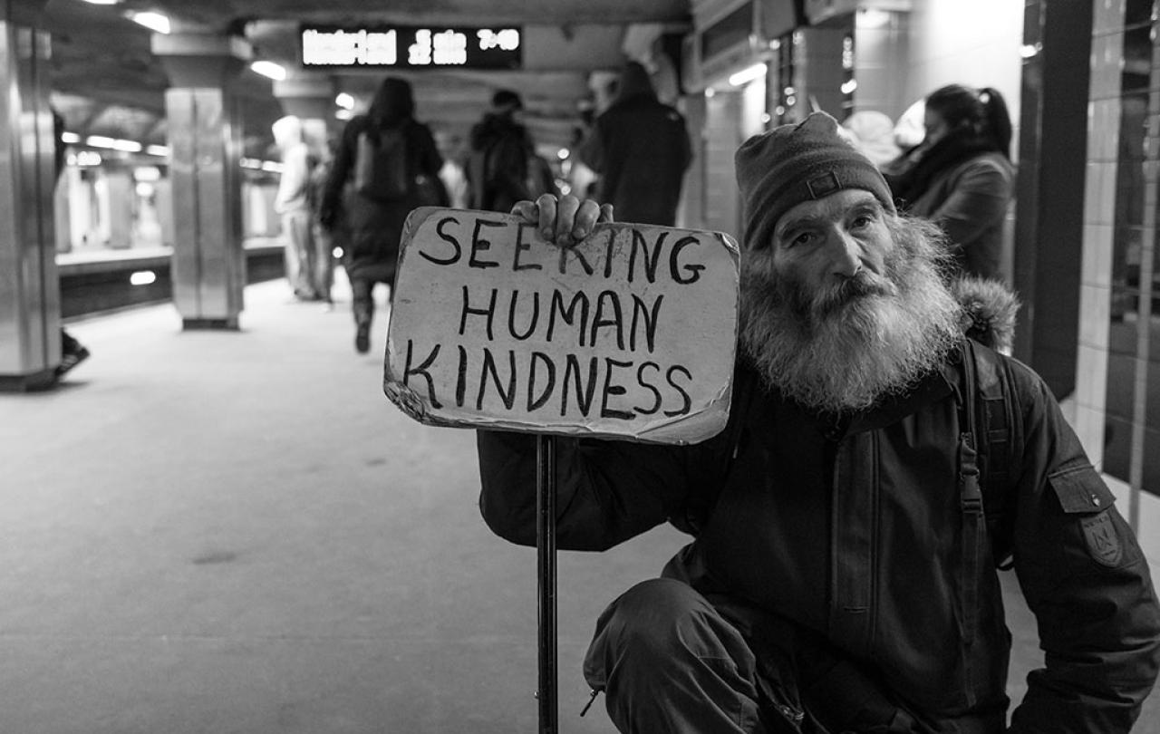 a man in a wheelchair sits in a subway station holding a sign reading 'seeking human kindness'.